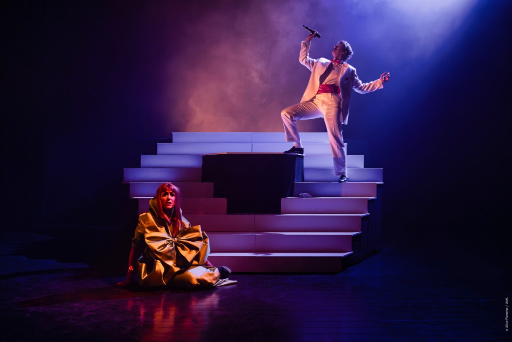 Une personne assise par terre en avant scène et une autre sur un escalier derrière en train de chanter. La lumière est tamisée type cabaret. L'escalier évoque certaines scénographies de programmes télé ou des shows.