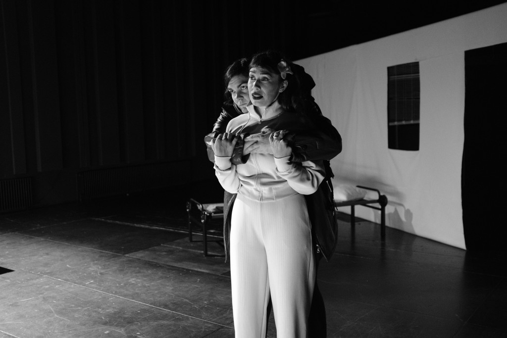 Deux femmes, celle devant habillée en blanc, celle derrière elle la serre dans ses bras. C'est une phot en noir et blanc.