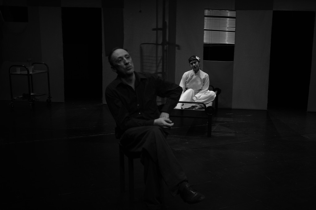 C'est une photo en noir et blanc. Sur le devant un personnage en chemise, une jambe croisée sur l'autre, comme s'il était en train de parler. Derrière lui asise sur un lit une personne habillée en blanc l'observe.