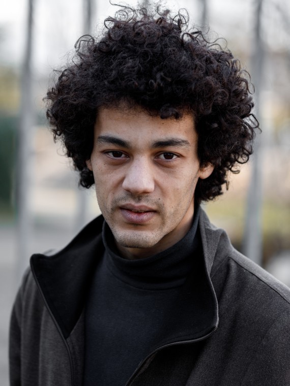 C'est le portrait de Ali Lamadli, il a les cheveux bouclés et il porte une veste marron foncé.