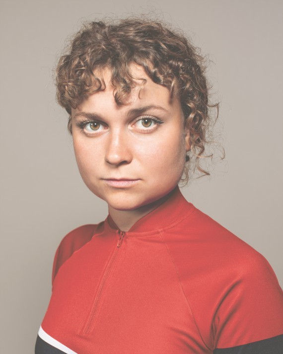 C'est le portrait de Emma Perez, elle porte un t-shirt orange elle a une frange bouclées.