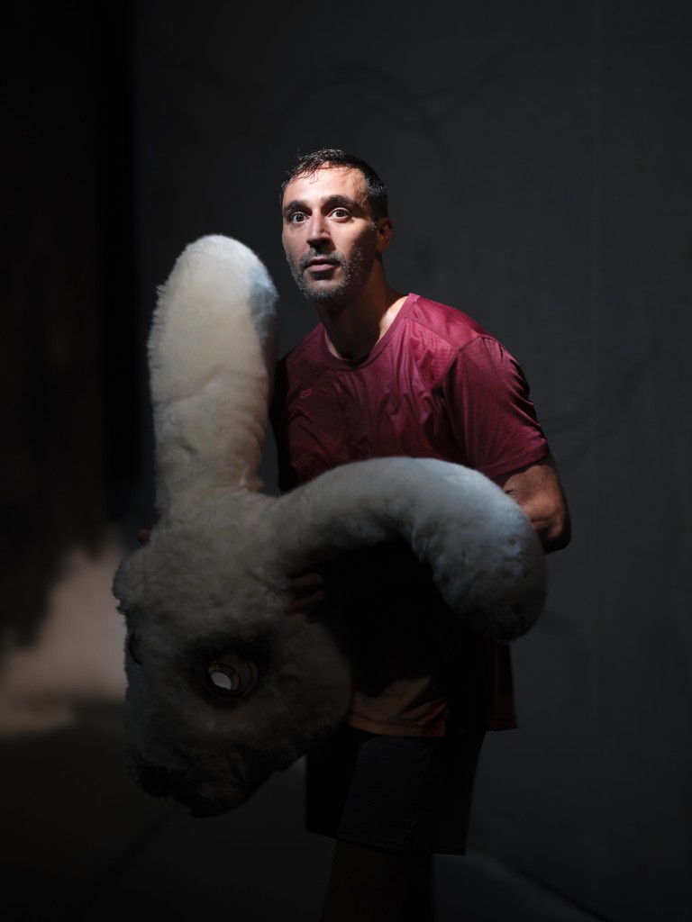 Une personne en train de tenir le masque dans lapin. Derriere elle de la fumée. La personne a l'air appeuré. Le masque c'est un gros masque comme celui des mascottes des stades.