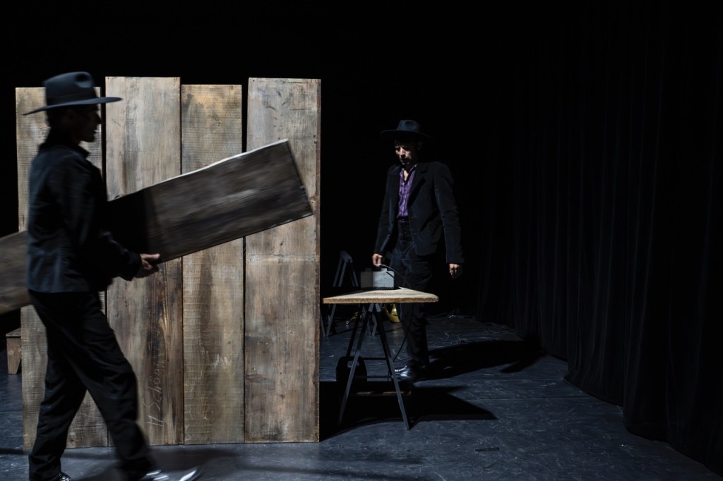 Une parroi en bois, à droite une table. Derriere la table une figure avec un chapeau noir. Devant la parroi une personne est en tran de transporter una palnche en bois.