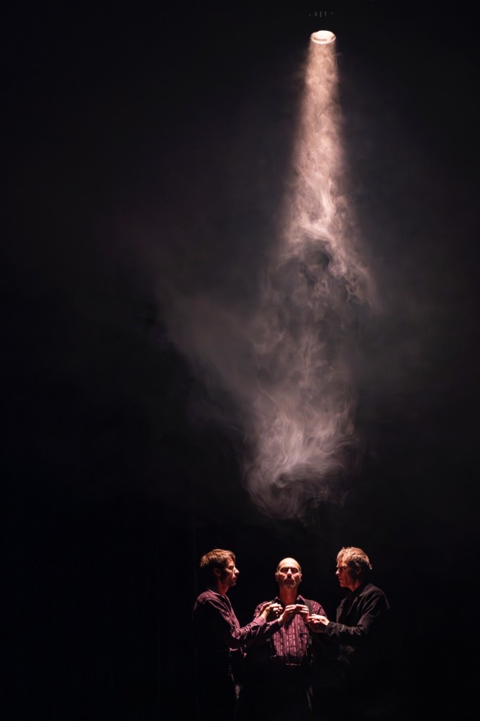 Trois personnes presque dans le noir. On dirait que ces personnes sont bien habillées. C'est comme si elles etaient en train de prier, une lumière venant du haut illumine un peu leurs visages.